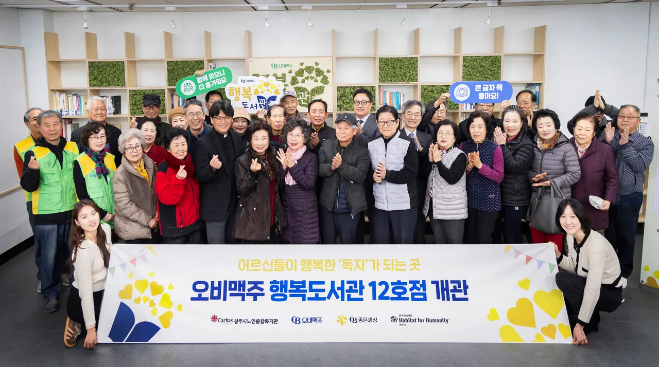 오비맥주 행복도서관 12호점 개관 행사에서 시니어들과 관계자들이 도서관 책장 앞에 모여 현수막을 들고 기념촬영을 하는 모습