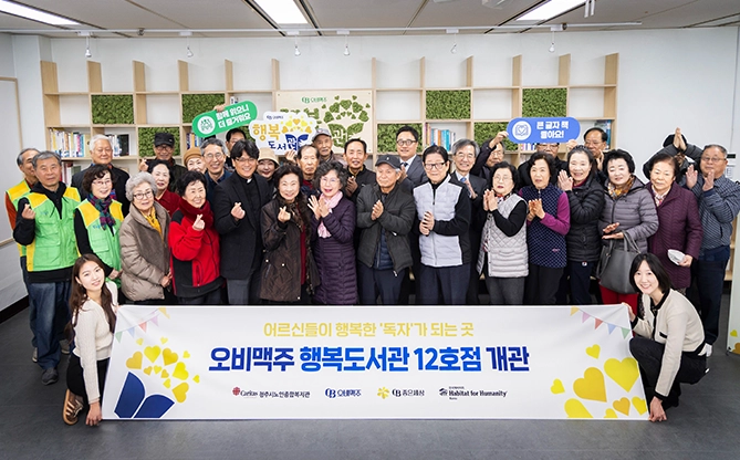 오비맥주 임직원과 도서관 내부에서 지역 어르신들과 ‘오비맥주 행복도서관 12호점 개관’ 현수막을 들고 함께 기념촬영하는 모습입니다.